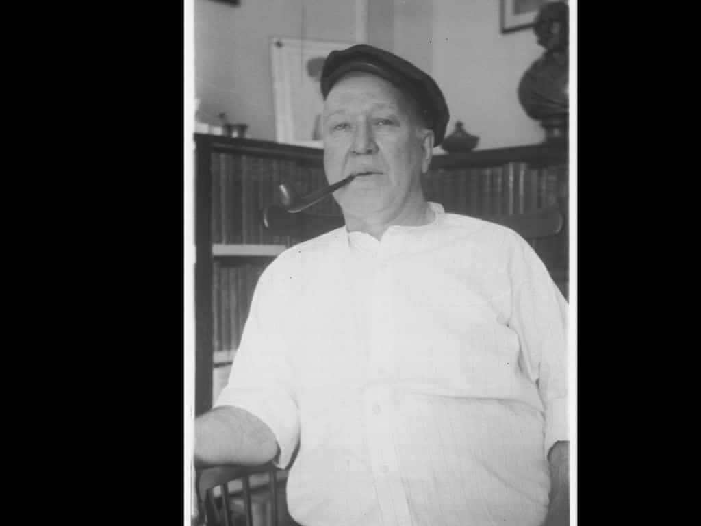 Black and white photo of Charles Gildersleeve wearing a white buttondown shirt and flat cap, and smoking a pipe. There are blurred shelves of books in the background.