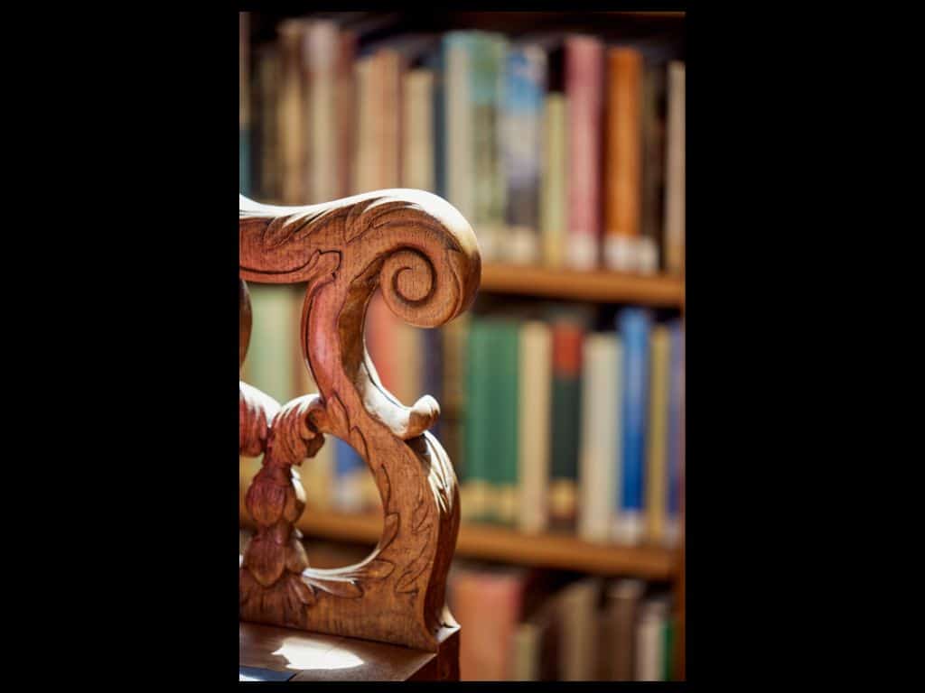 Close-up of an ornate wooden chair arm with carved scrollwork, set against the blurred backdrop of library bookshelves.