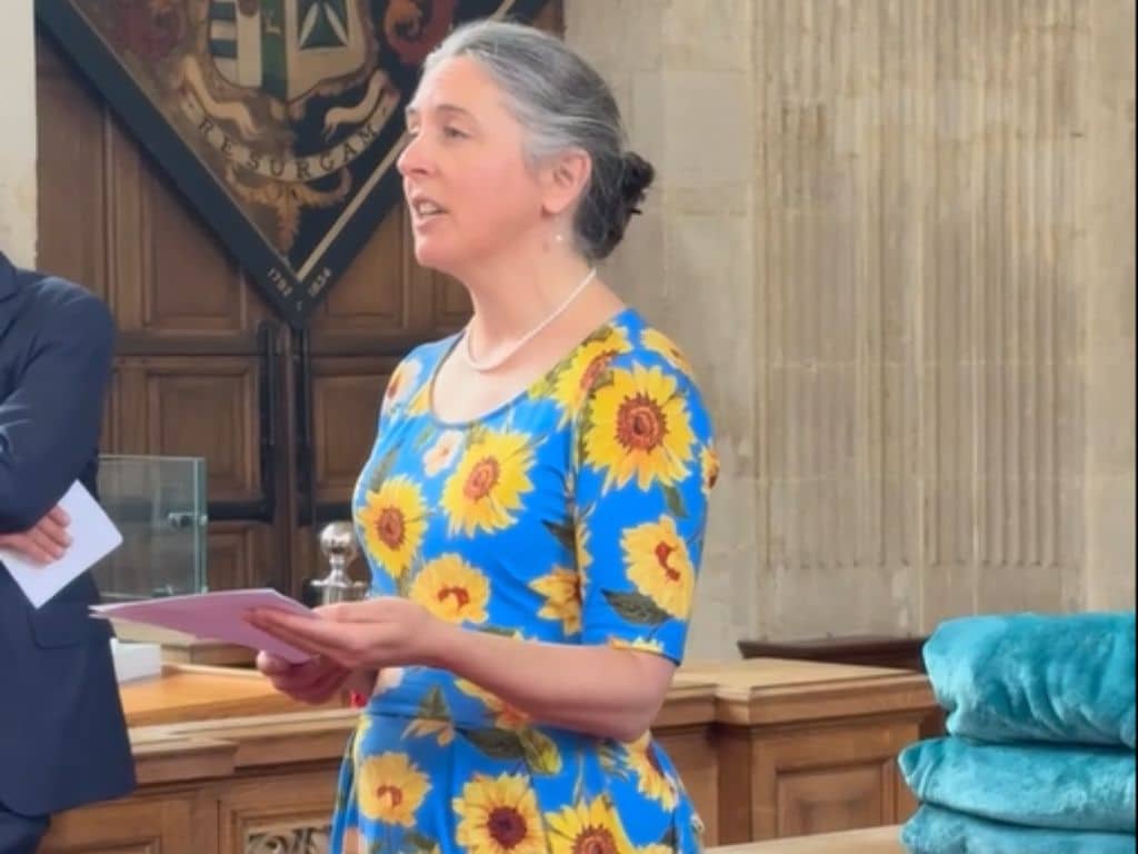 Lucy Matheson in a blue dress with sunflowers addressing attendees in the College library