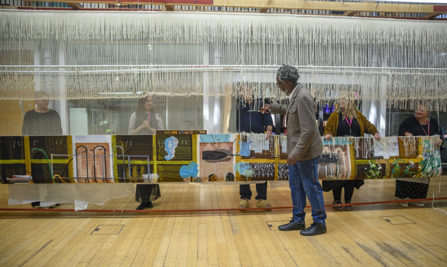 A group of weavers and Hurvin Anderson holding up a newly completed tapestry still attached to the loom, its vibrant patterns of barbershop interiors stretching across the frame.