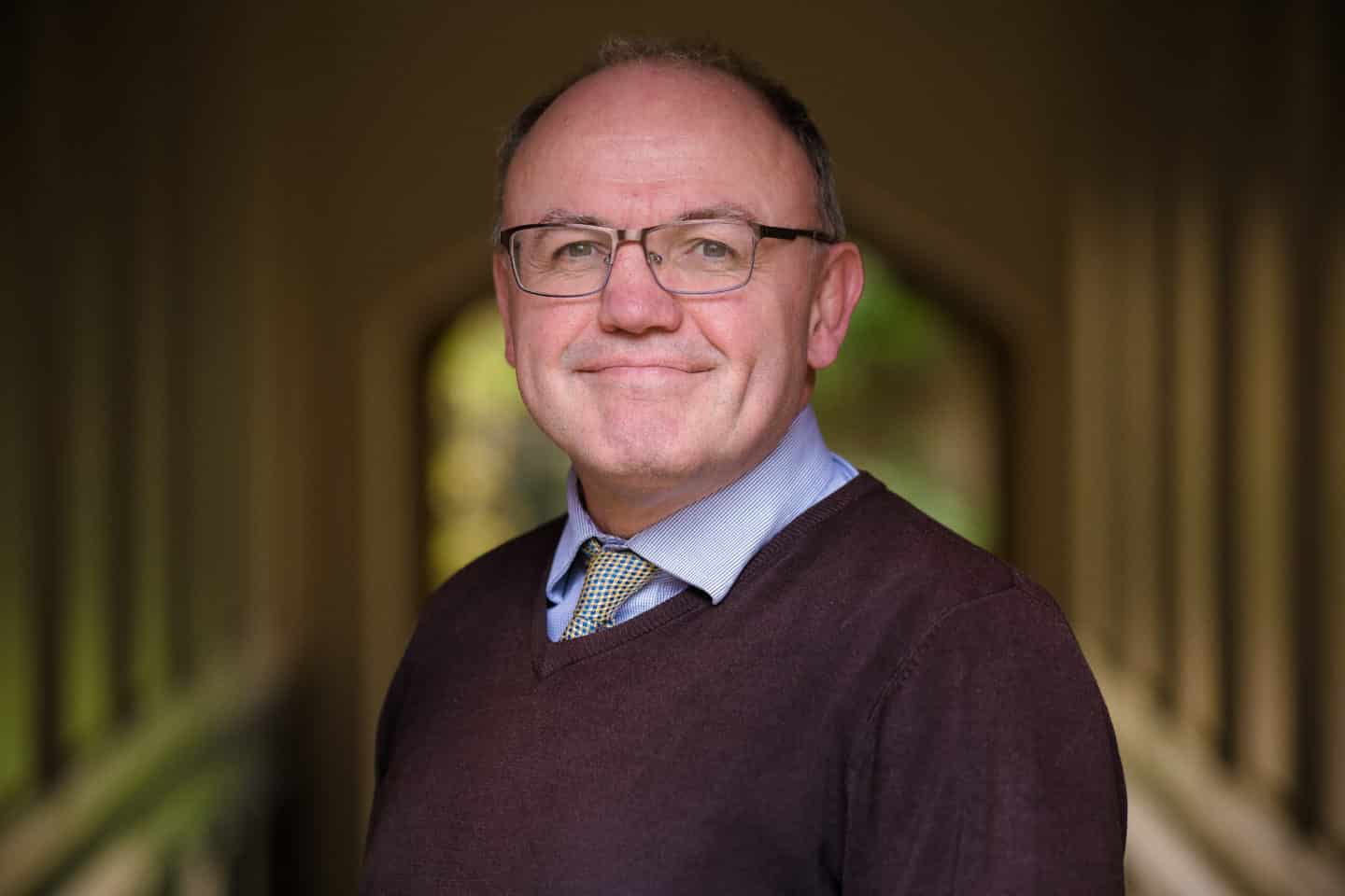 Headshot of Dr Perry Gauci, a man in glasses, a shirt and tie and a brown jumper