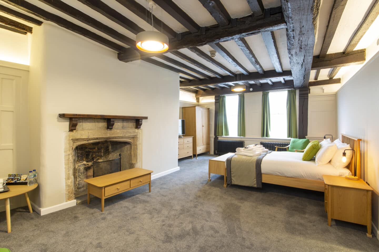 Bedroom with wooden beams on the ceiling, a double bed with pillows, cushions, towels and toiletries, a bedside table with lamp, a fireplace, a coffee table with a selection of tea, coffee, water and biscuits, a chest of drawers and wardrobe, a monitor, two radiators, and two large windows with green curtains.