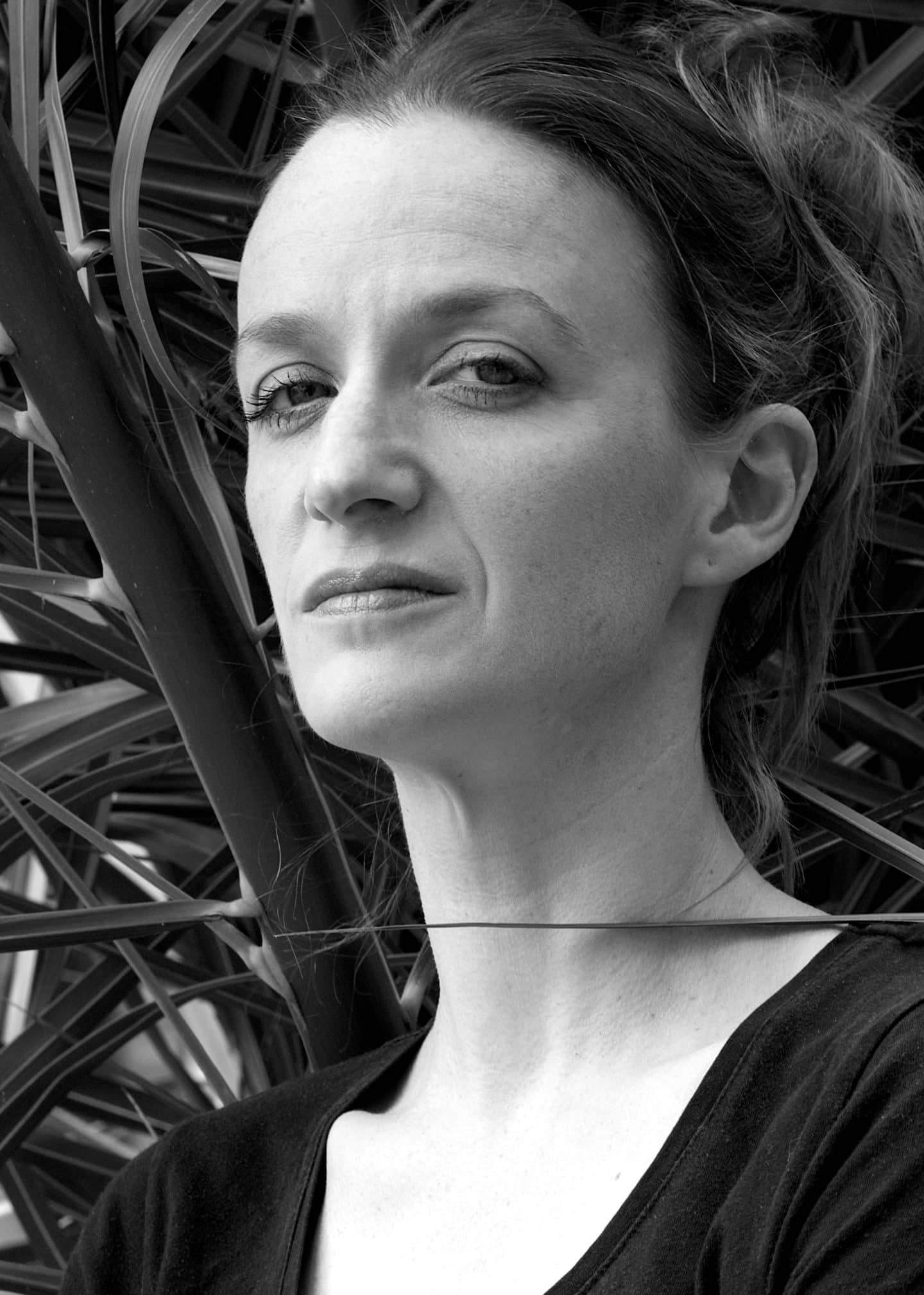 Greyscale photograph of Kate Smurthwaite, a woman standing before a mass of thin leaves