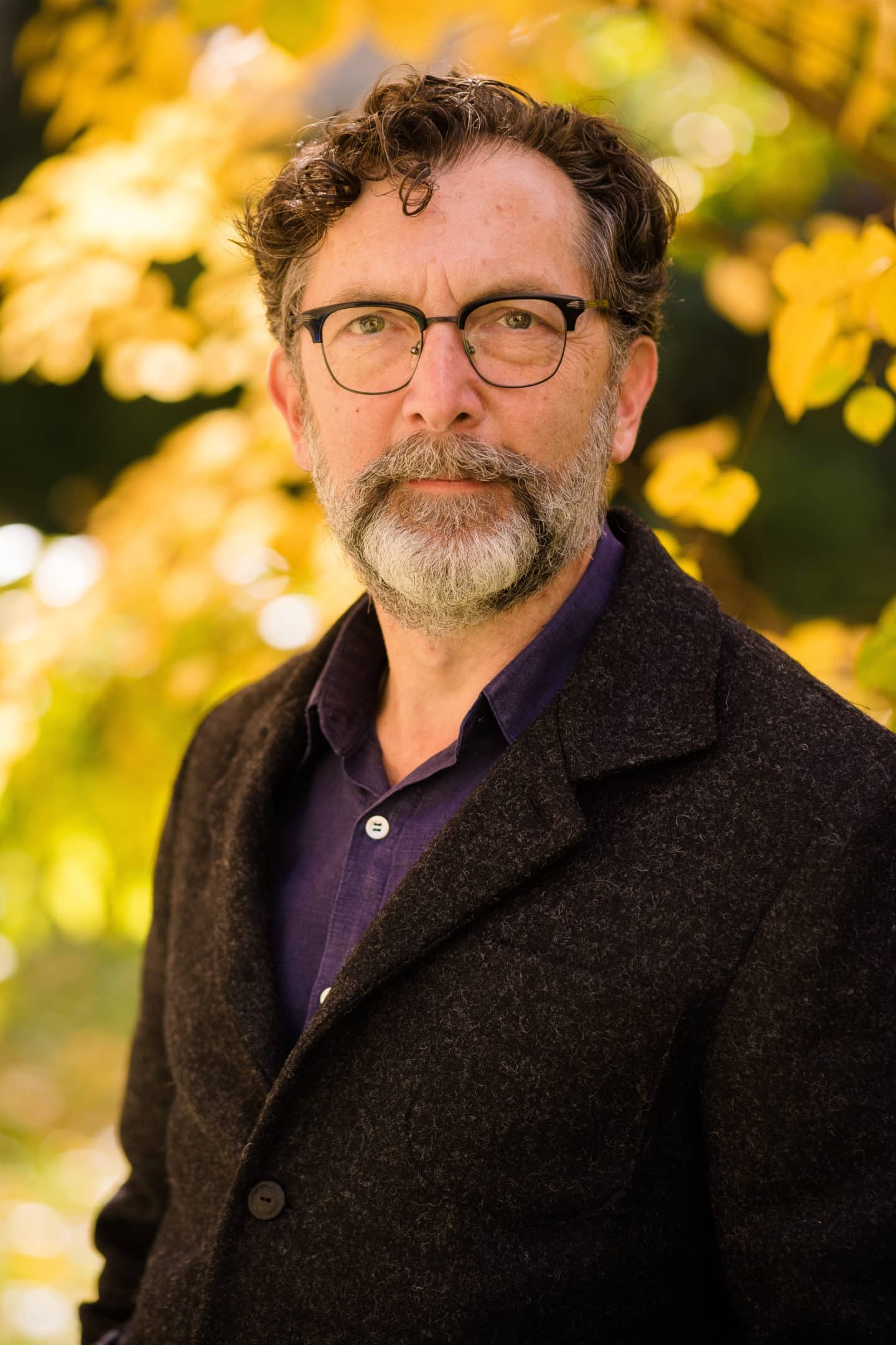 Headshot of Peter McCollough, a man in a purple shirt, a black coat, and glasses