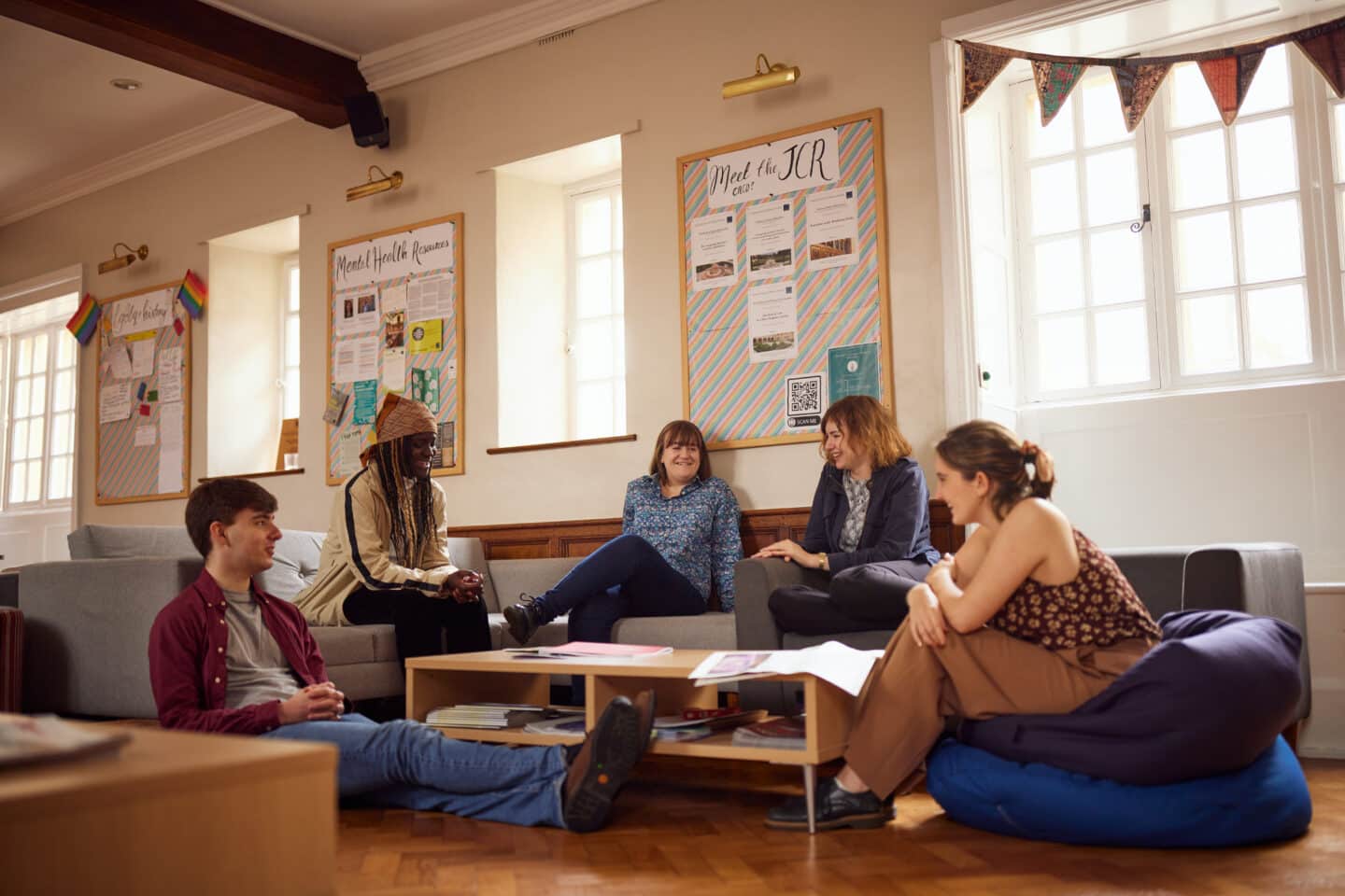 Five smiling students sitting around a table in the Lincoln College Junior Common Room. On the walls in the background are three boards, respectively titled "Meet the JCR', 'Mental Health Resources' and 'lgbtq+ history'