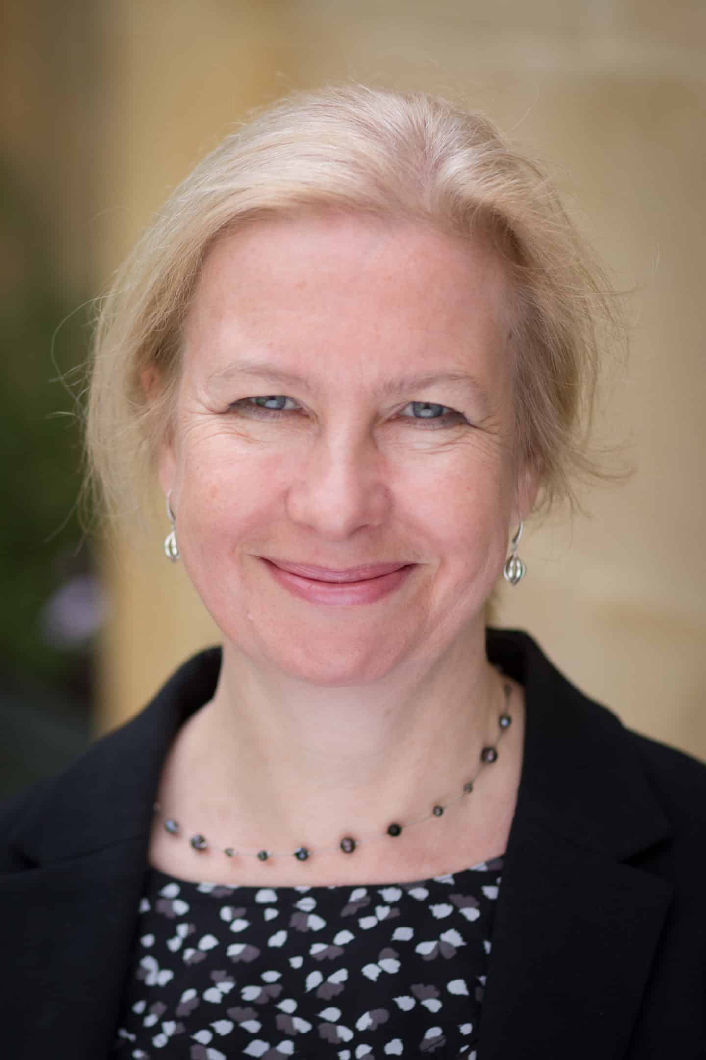 Headshot of Susan Harrison, a woman in a black patterned top, black jacket, necklace and earrings