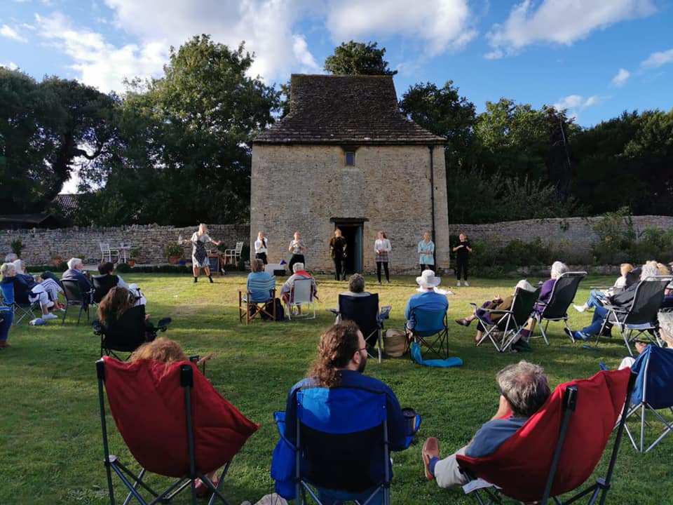 An outdoor theatre performance taking place in front of a historic stone building with a thatched roof. The audience, seated in foldable chairs on a grassy lawn, watches as actors perform in the open-air setting. The sky is blue with scattered clouds, and the surrounding area is enclosed by a stone wall with trees in the background. The performers are dressed in period-style clothing, engaging in a dramatic scene.