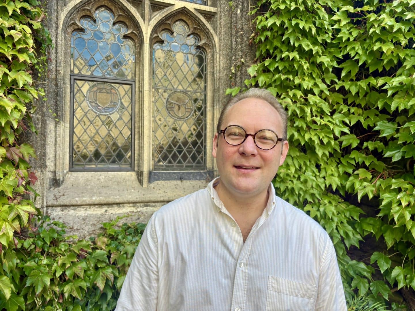 Oliver Walker in a white shirt standing in front of stained glass window surrounded by ivy
