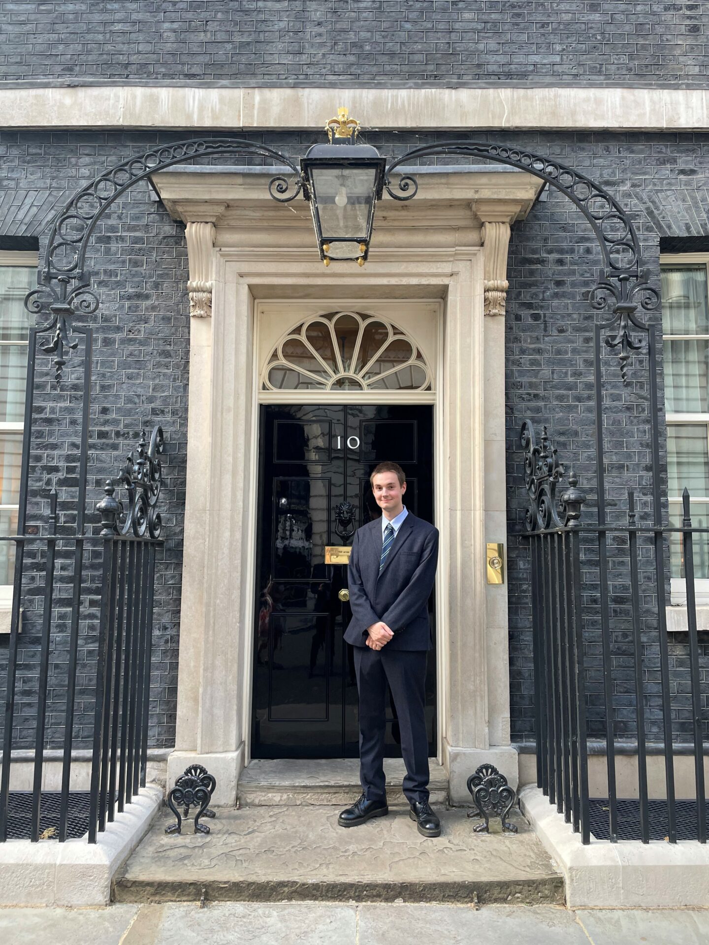 Headshot of Jacob Kelly, a young man in a suit standing in front of the door of 10 Downing Street